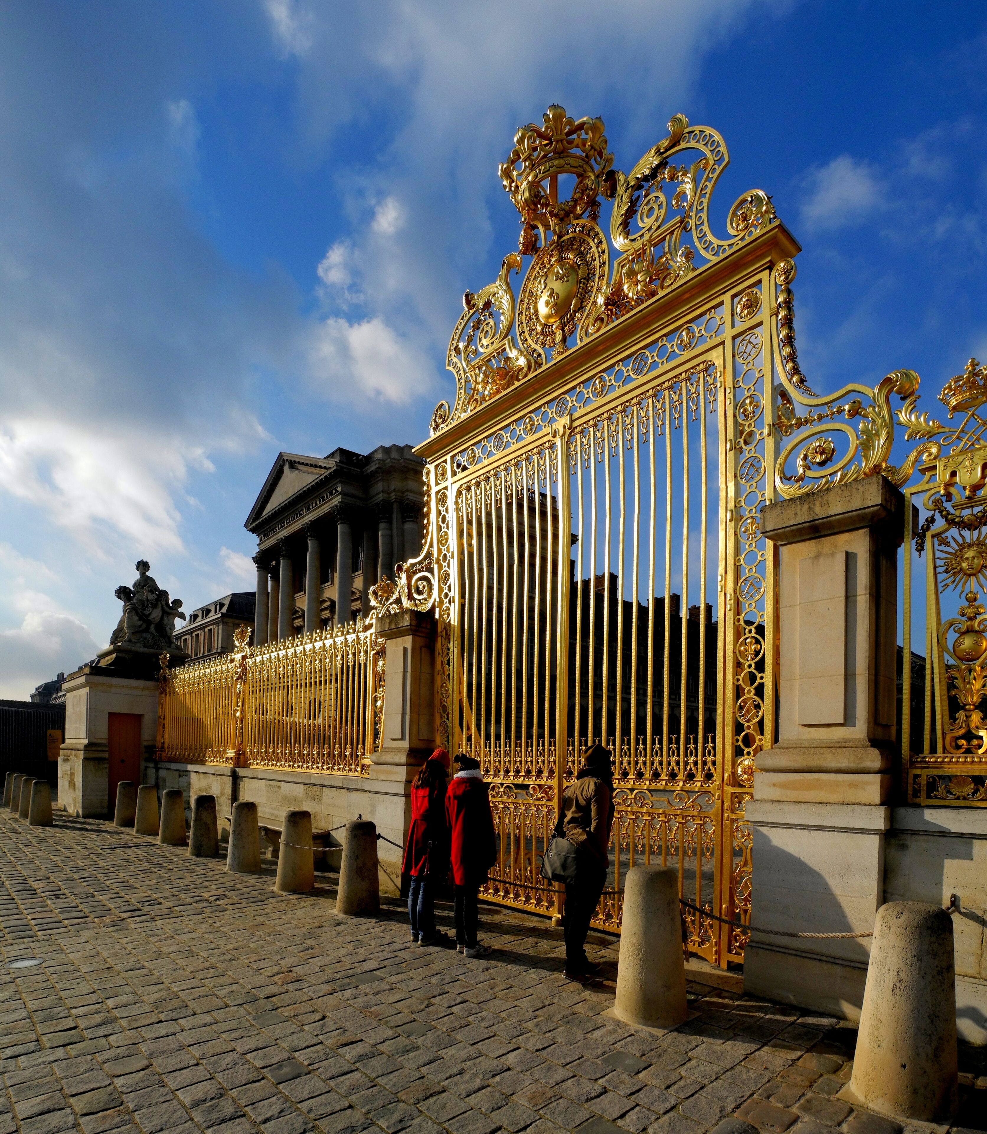 A grand golden gate with intricate designs, flanked by classical columns and a statue, set against a clear blue sky.
