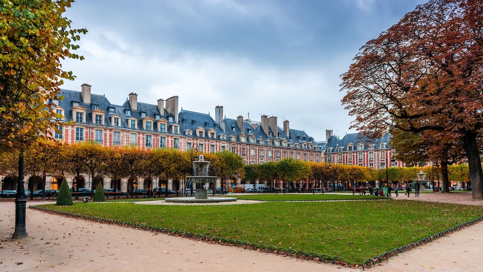 Un edificio histórico con fachadas rojas y blancas, una fuente central y un parque bien cuidado con árboles y césped.