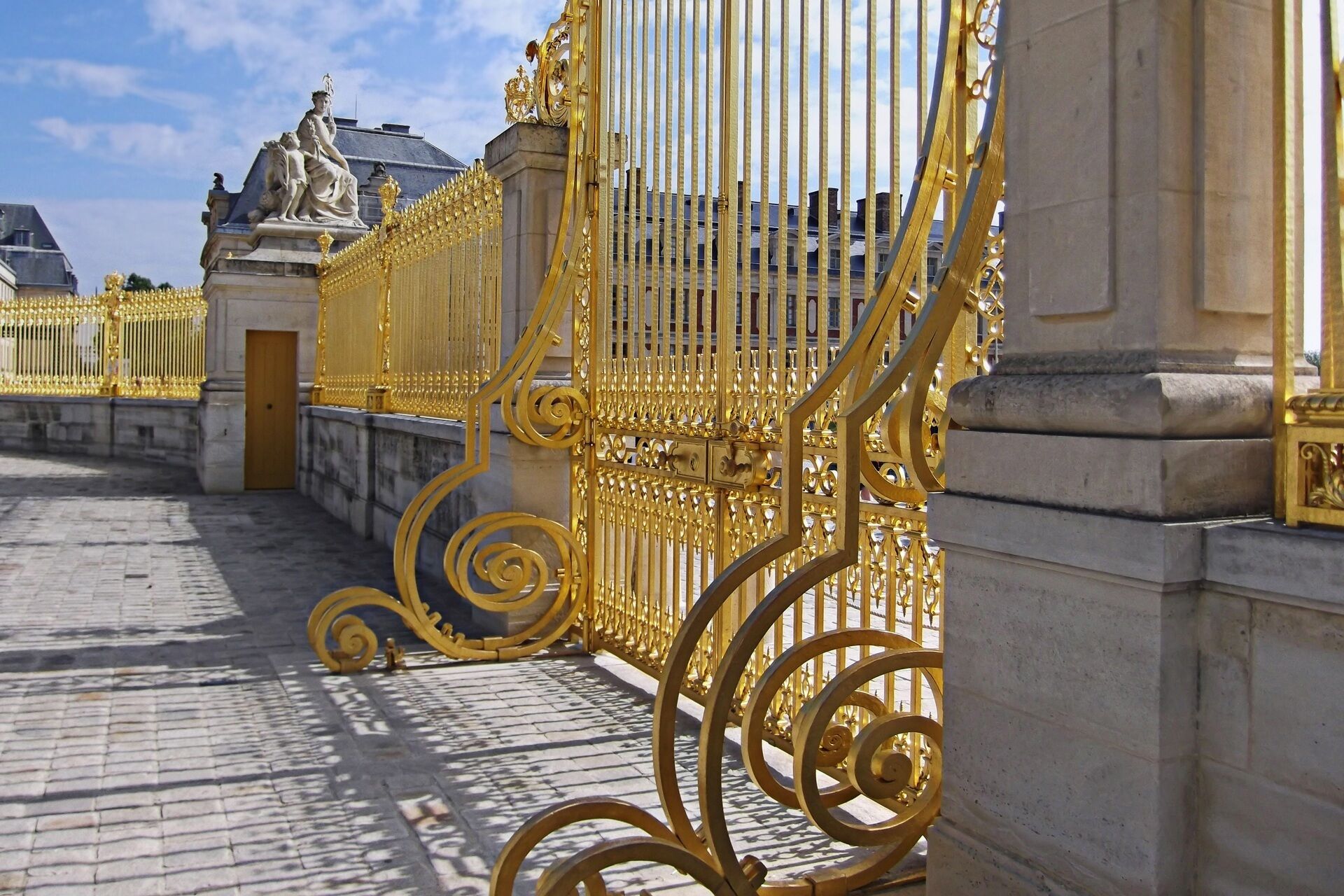 A golden gate with intricate designs, leading to a building with statues and a domed roof.