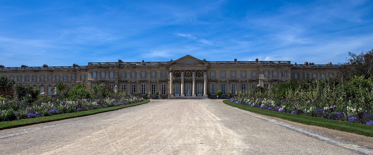 A large classical building with a symmetrical facade, a central entrance, and a gravel driveway leading to it.