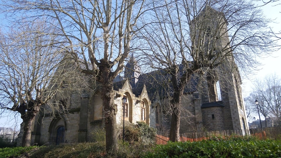A historic stone church with arched windows and a tower, surrounded by leafless trees and parked cars.