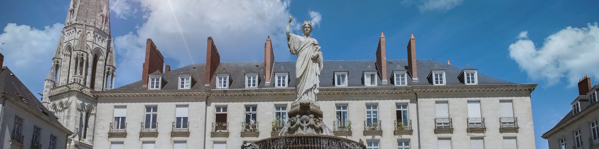 Ein historisches Gebäude mit einer Statue einer Frau, die ein Kind hält, einem Brunnen mit mehreren Figuren und einem hohen Turm.