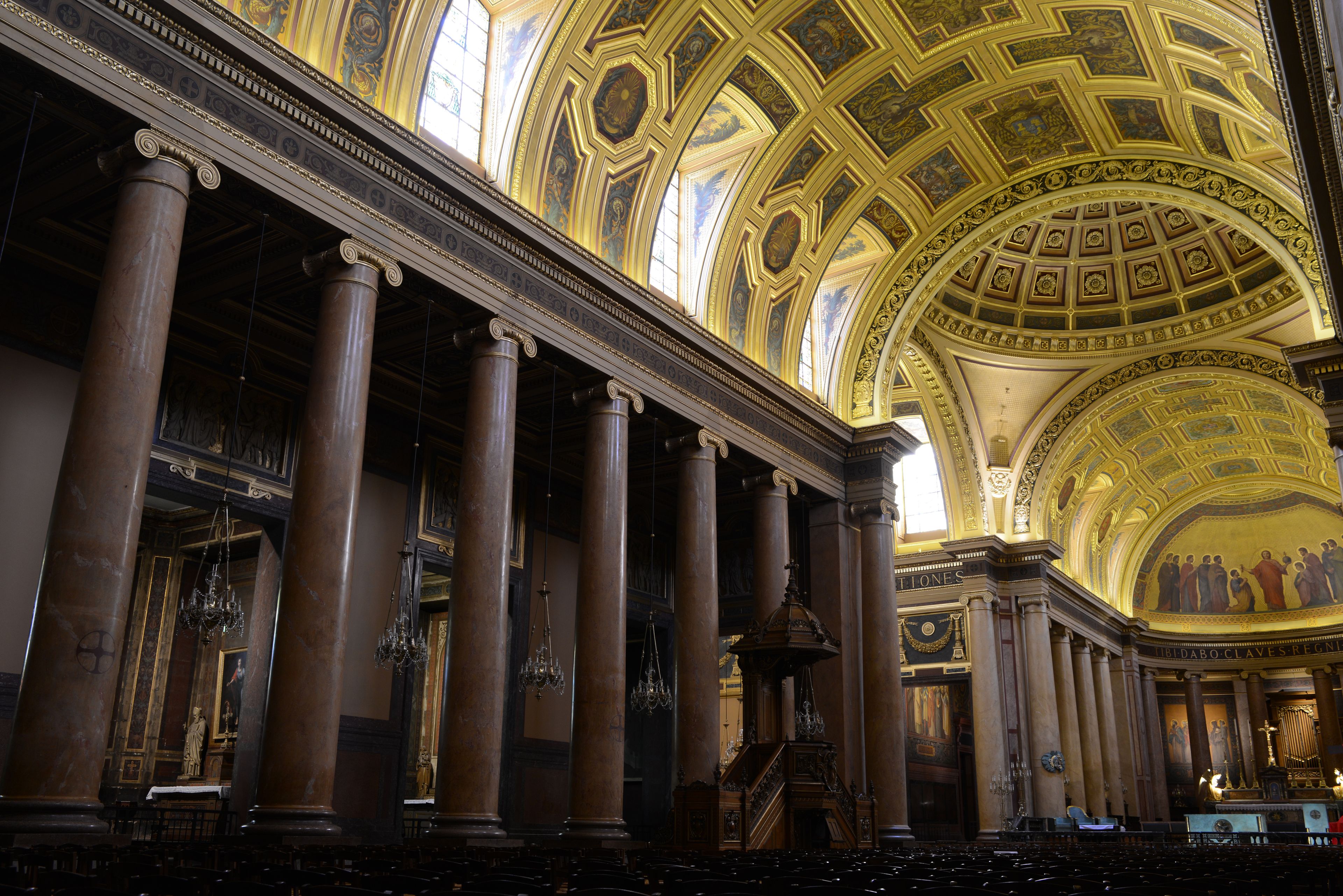 A grand hall with ornate columns, a high vaulted ceiling with intricate designs, and a large chandelier.