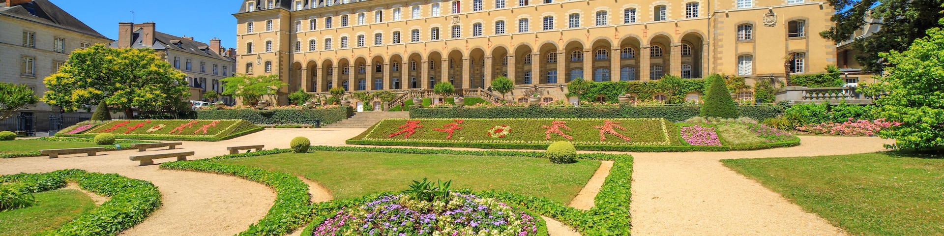 Un bâtiment historique à la façade symétrique, doté d’une cour centrale avec un parterre floral circulaire et d’un jardin à la française aux motifs géométriques.