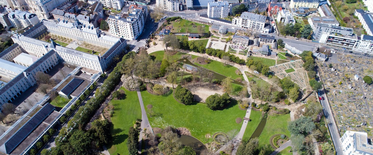 An aerial view of a park with pathways, a pond, and surrounding buildings.