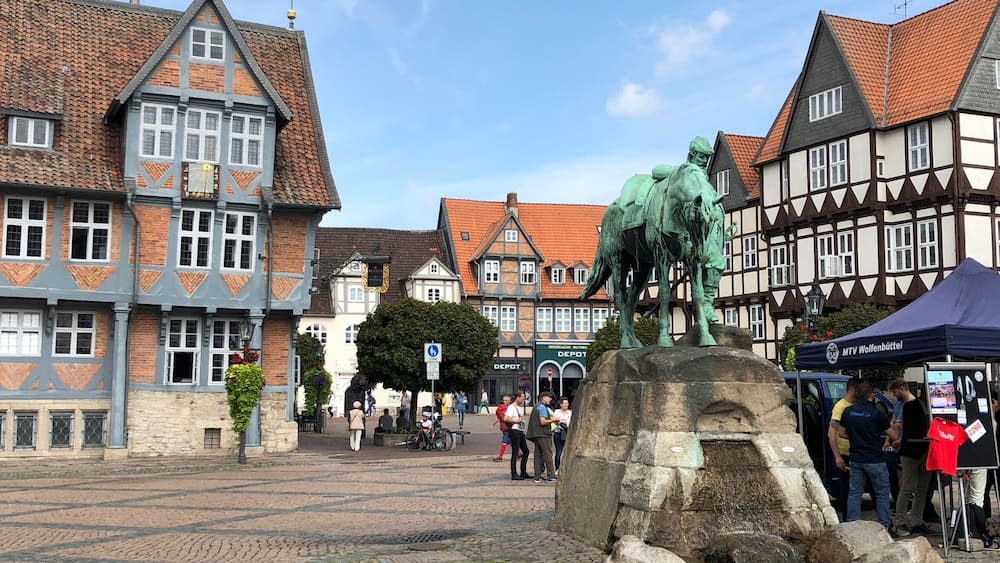 Ein Kopfsteinpflasterplatz mit traditionellen Fachwerkhäusern, einer Pferdestatue und einem Zelt mit der Aufschrift 'MTV Wolfenbüttel'.