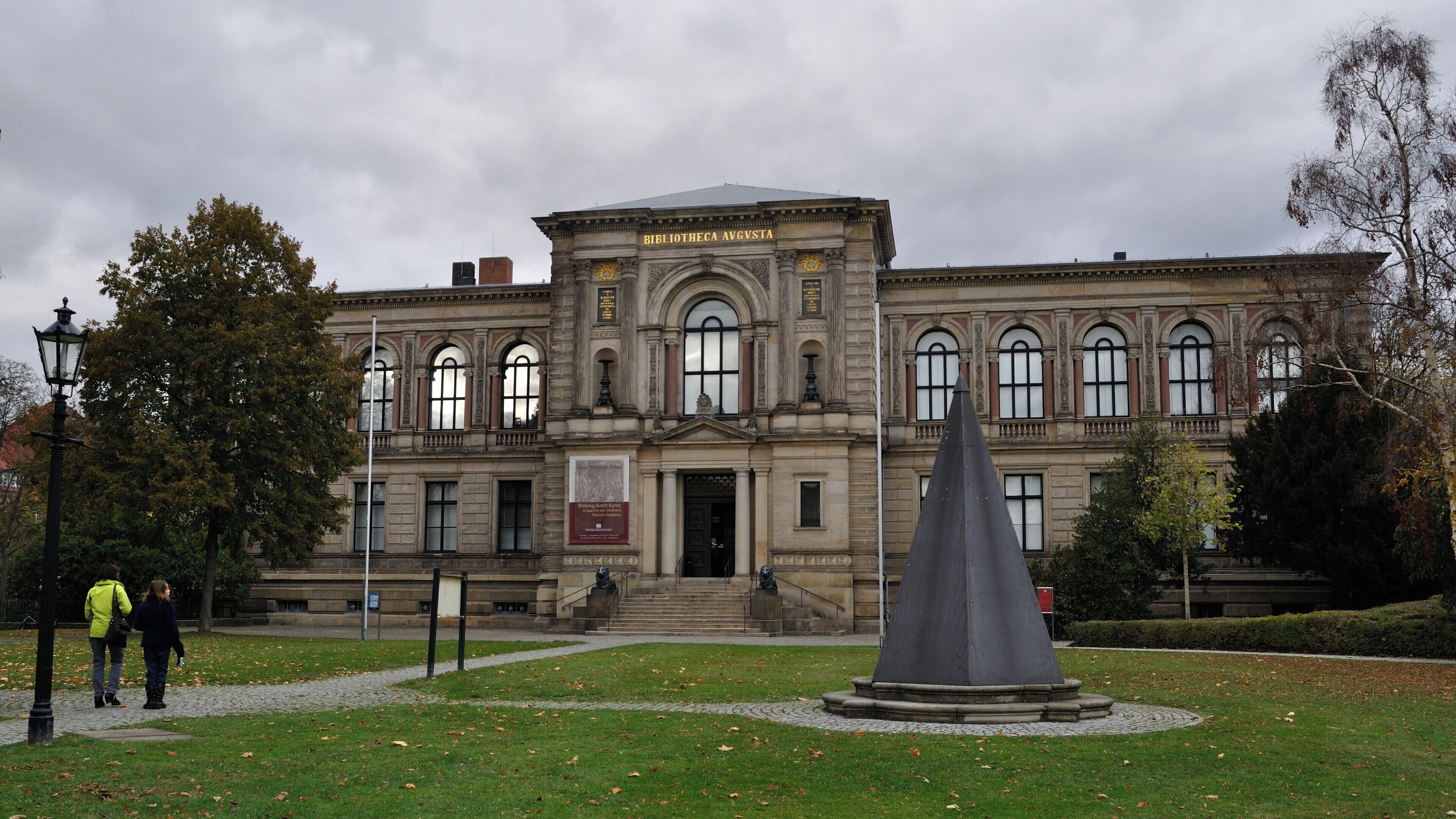 A historic building with a prominent entrance, a statue in front, and a well-maintained lawn.
