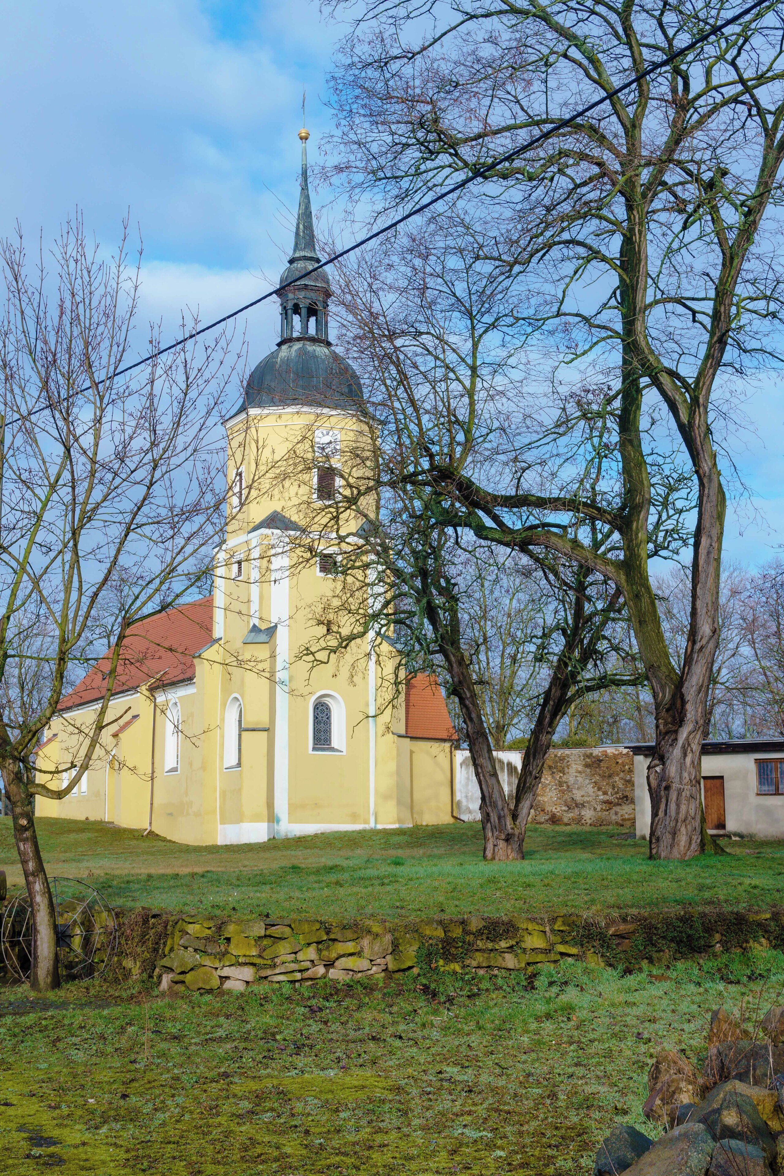 A church with a tall bell tower and a dome, surrounded by trees and a stone wall.