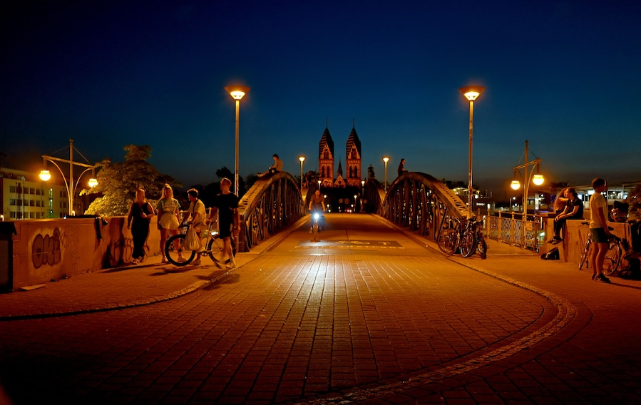 A bridge at night with street lamps, a gothic church in the background, and people walking and cycling.