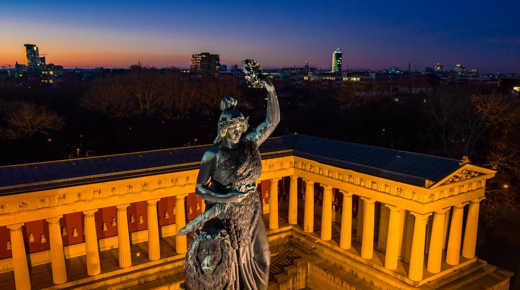 Una estatua de una mujer sosteniendo una corona de laurel, sobre un pedestal frente a un edificio clásico con columnas.