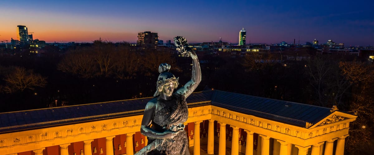 A statue of a woman holding a laurel wreath, standing on a pedestal in front of a classical building with columns.