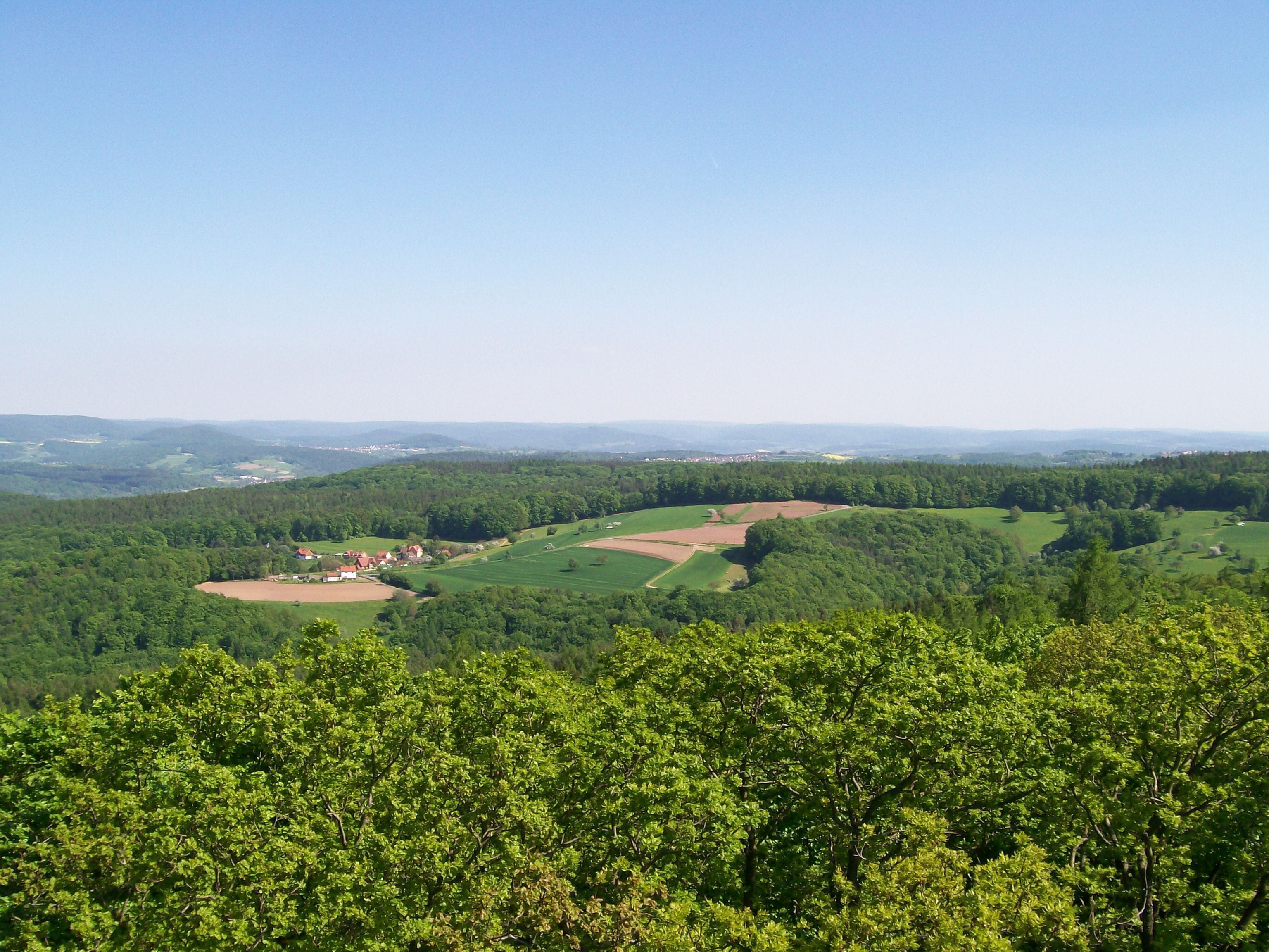 A lush green landscape with rolling hills, a small village, and distant mountains under a clear sky.