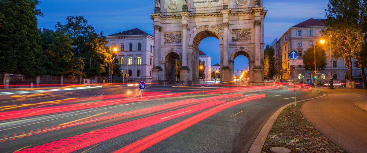 Ein historisches Tor mit Inschriften, umgeben von Bäumen und Gebäuden, mit Lichtspuren von Fahrzeugen auf der Straße.