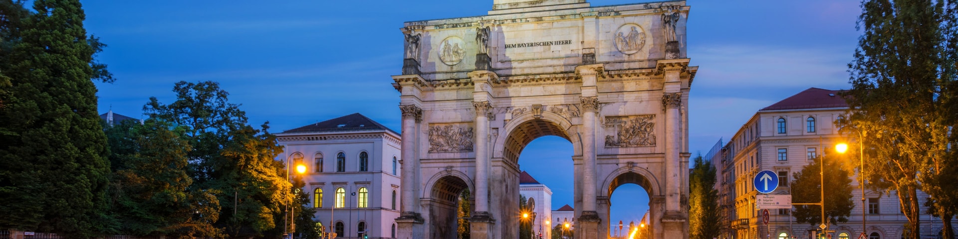 A historic archway with inscriptions, surrounded by trees and buildings, with light trails from vehicles on the road.
