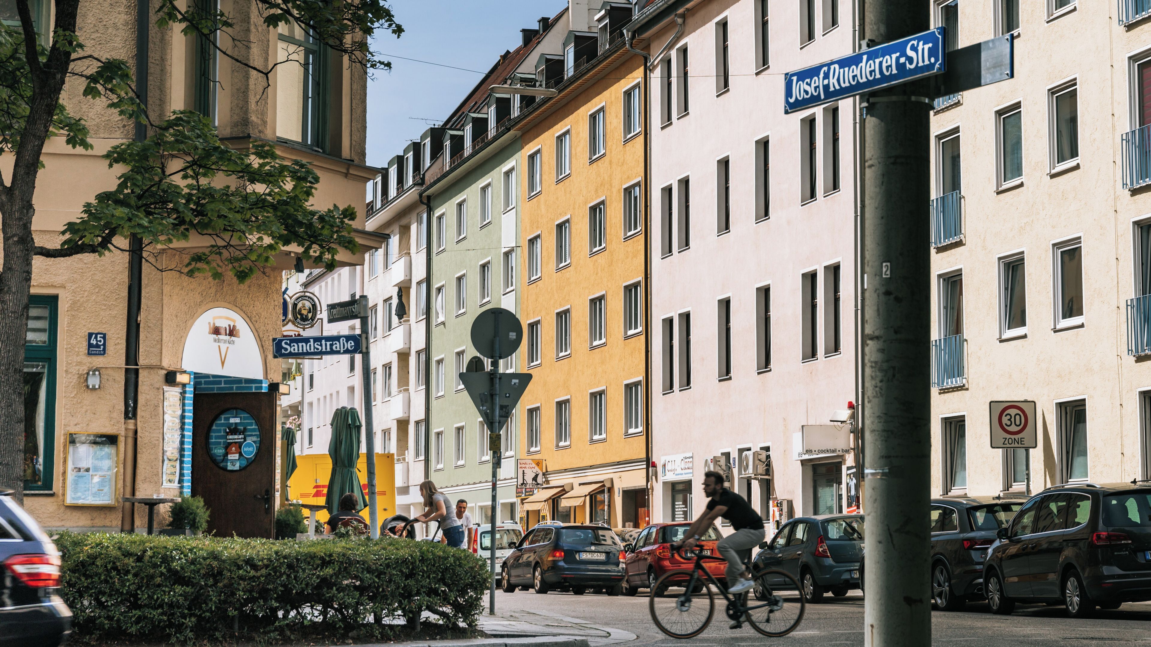 A street with buildings, cars, and a cyclist. Street signs are visible, including 'Josef-Ruederer-Str.' and 'Sandstrasse.'