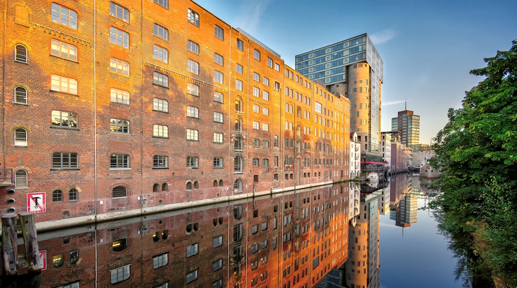 A river reflecting buildings, including a brick structure with arched windows and a modern glass building.