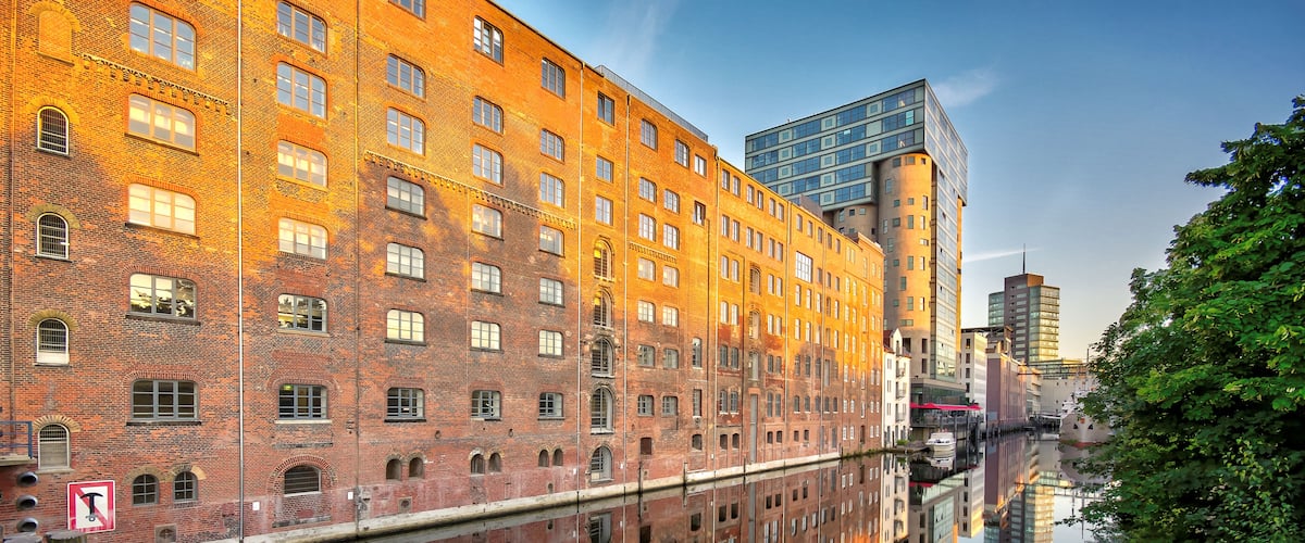 Ein Fluss, der Gebäude spiegelt, darunter ein Backsteinhaus mit Bogenfenstern und ein modernes Glasgebäude.