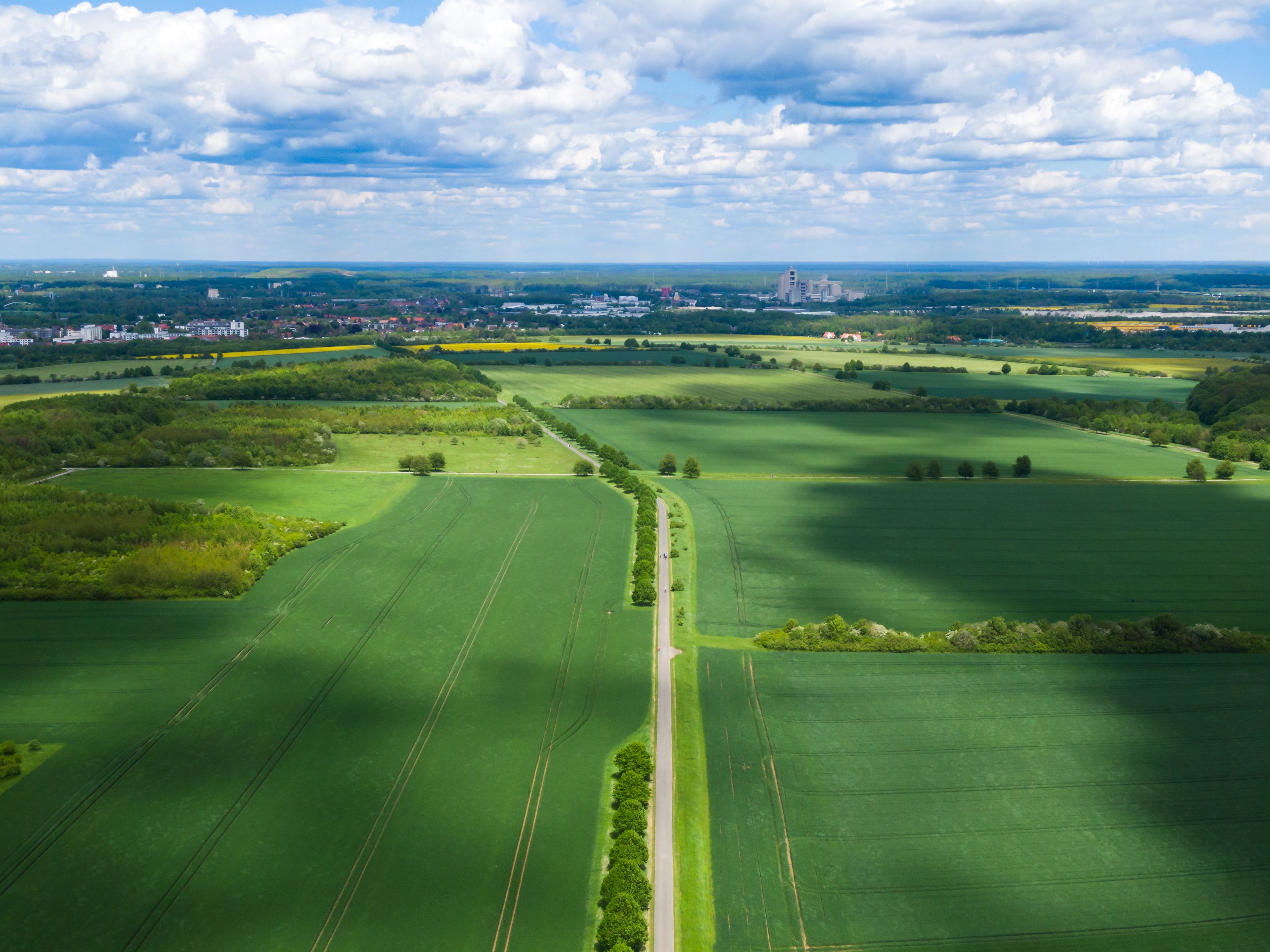 A vast green agricultural landscape with a winding road, distant buildings, and a cloudy sky.