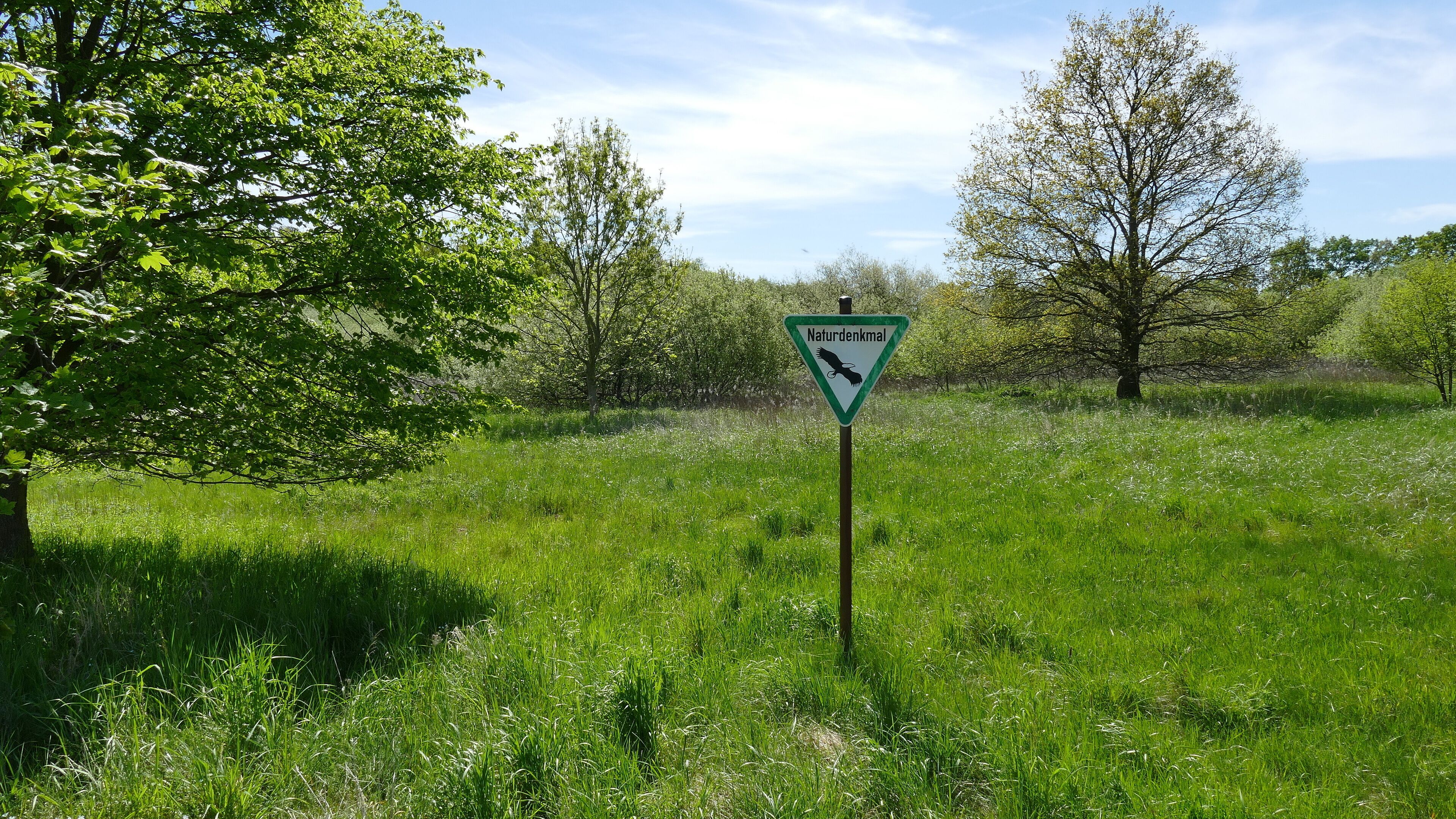 A sign with a bird symbol in a grassy field with trees.