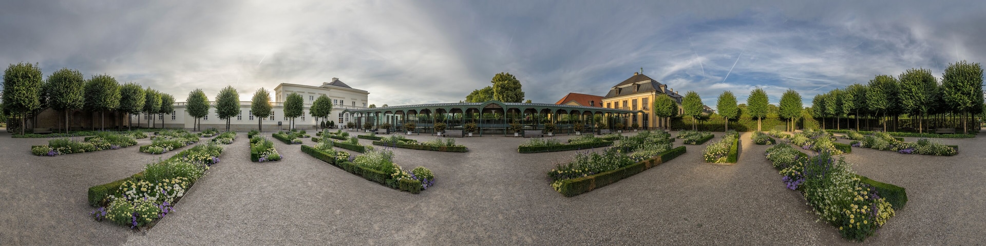Um jardim formal com canteiros florais simétricos, um pavilhão de vidro e arquitetura clássica.