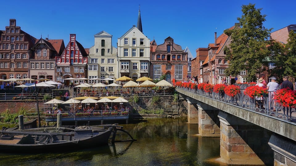 Eine Stadt am Flussufer mit historischen Gebäuden, einer Brücke mit roten Blumen und Booten, die an der Uferlinie liegen.