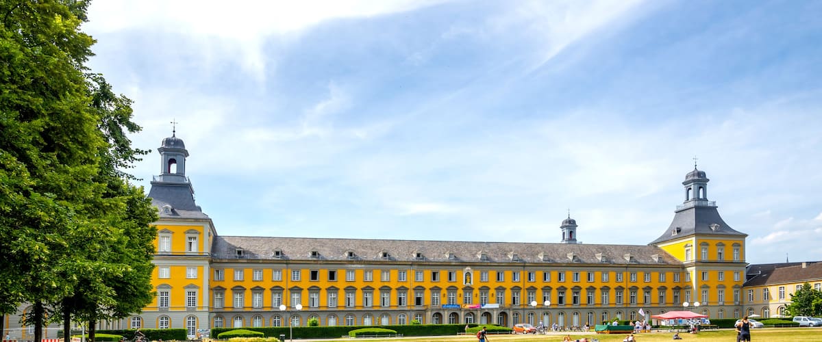 Ein großes gelbes Gebäude mit einem Glockenturm, umgeben von einem grünen Innenhof mit sitzenden und sich bewegenden Personen.