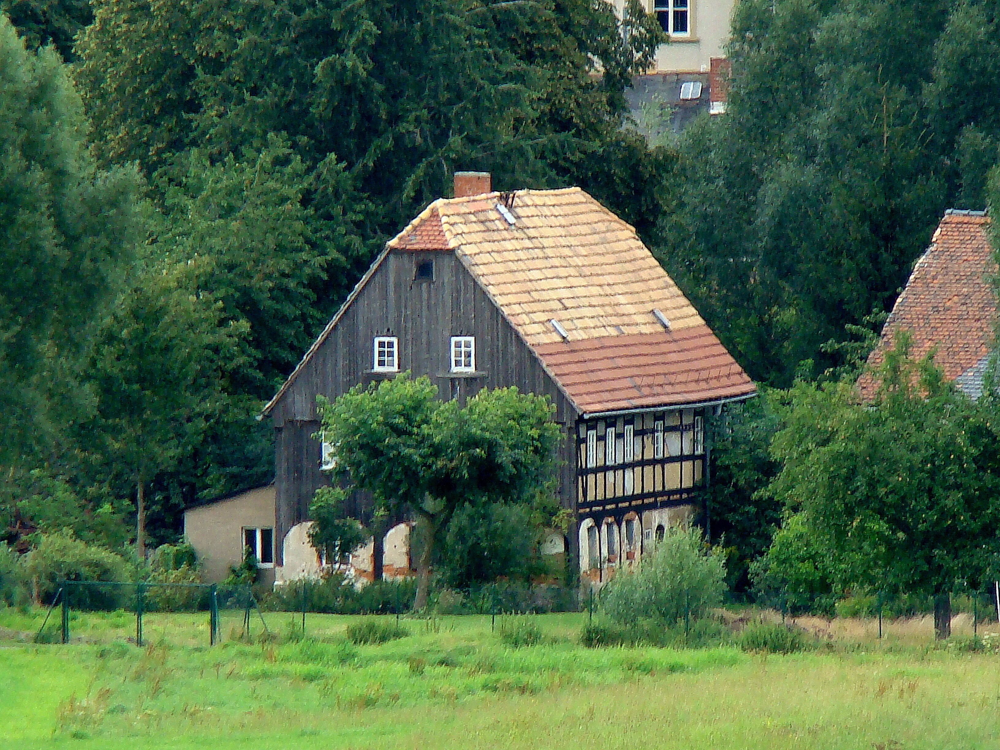 A traditional half-timbered house with a sloped roof, surrounded by lush greenery and trees.