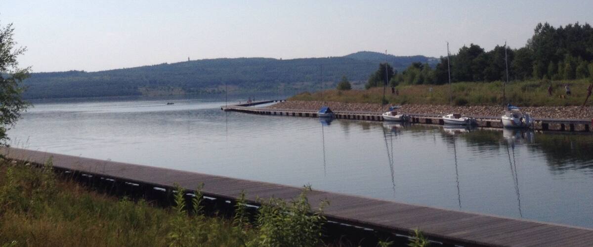 A calm lake with boats docked at a pier, surrounded by hills and greenery.