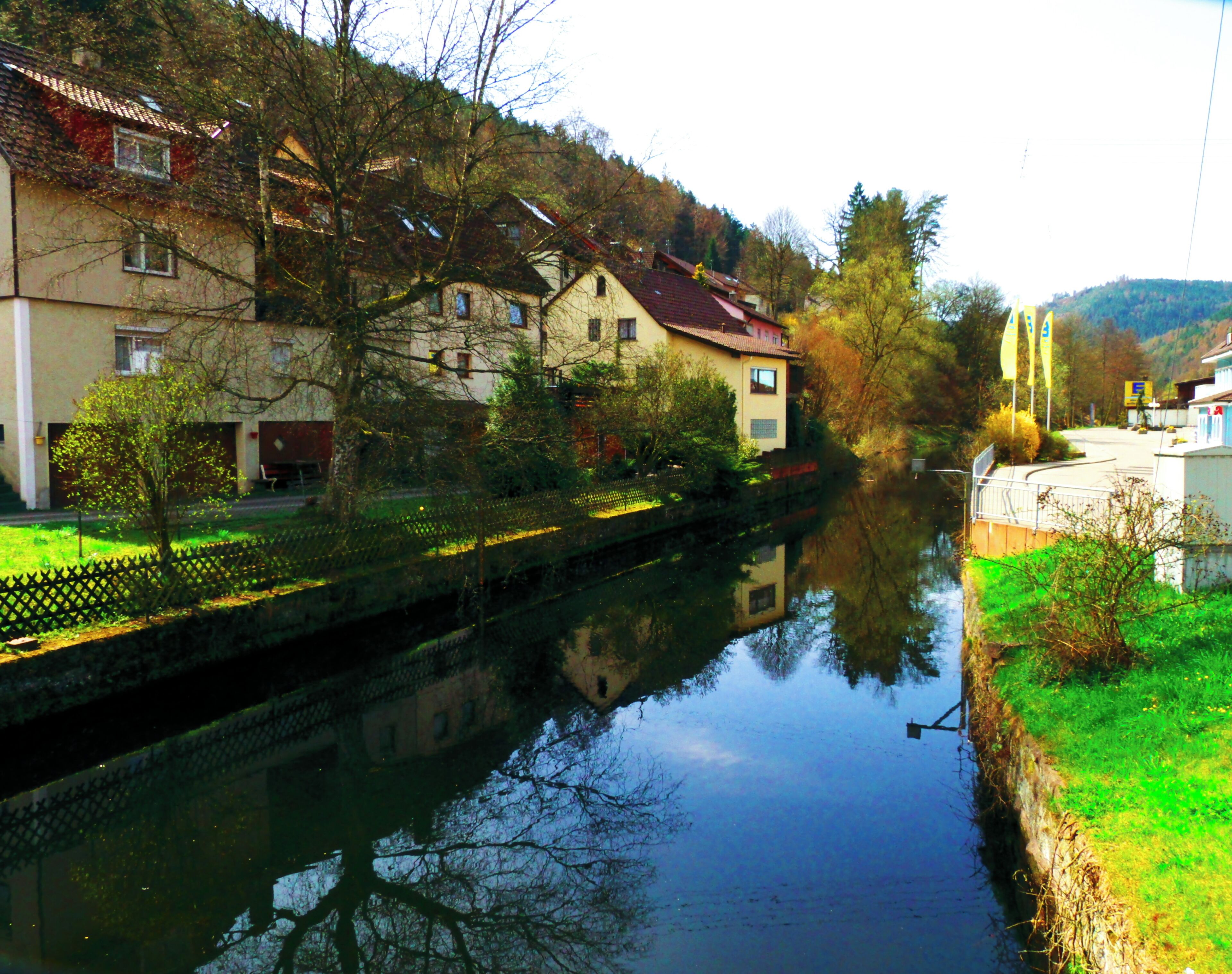 A calm river reflecting buildings and trees, surrounded by greenery and hills.