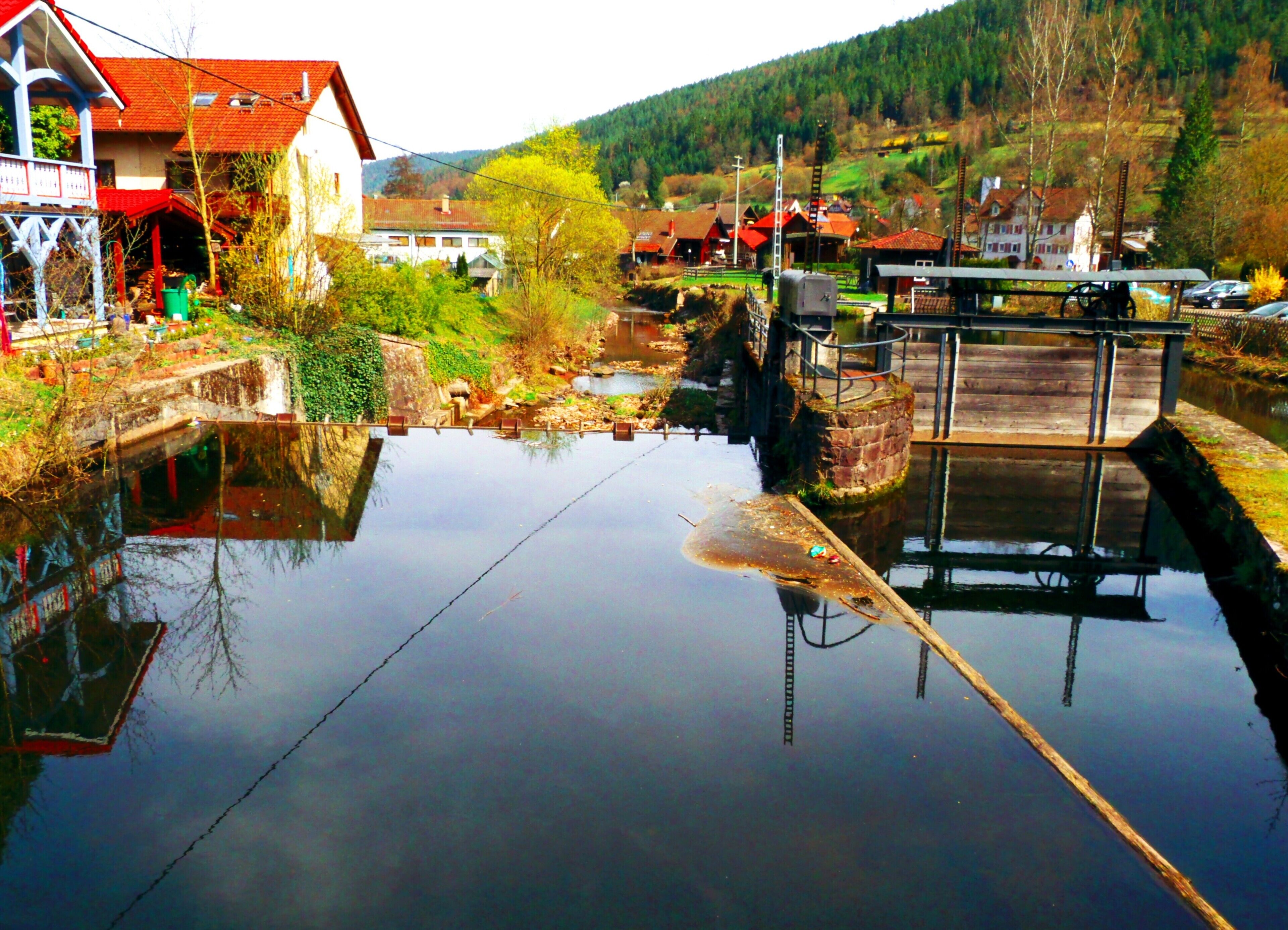 A calm river with a wooden dock, houses, and a forested hillside in the background.
