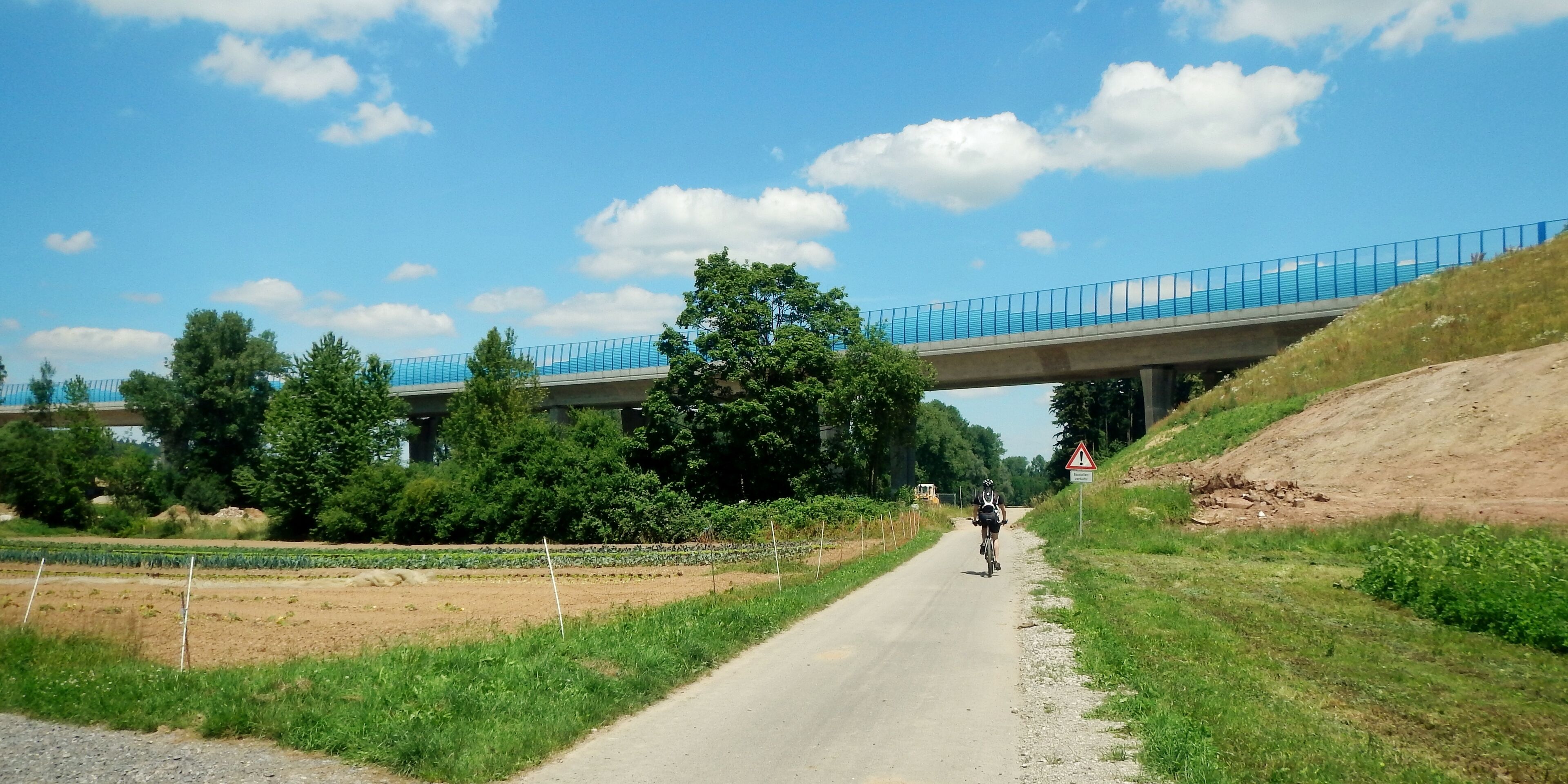 A cyclist on a paved road under a blue bridge with a warning sign, surrounded by greenery and fields.