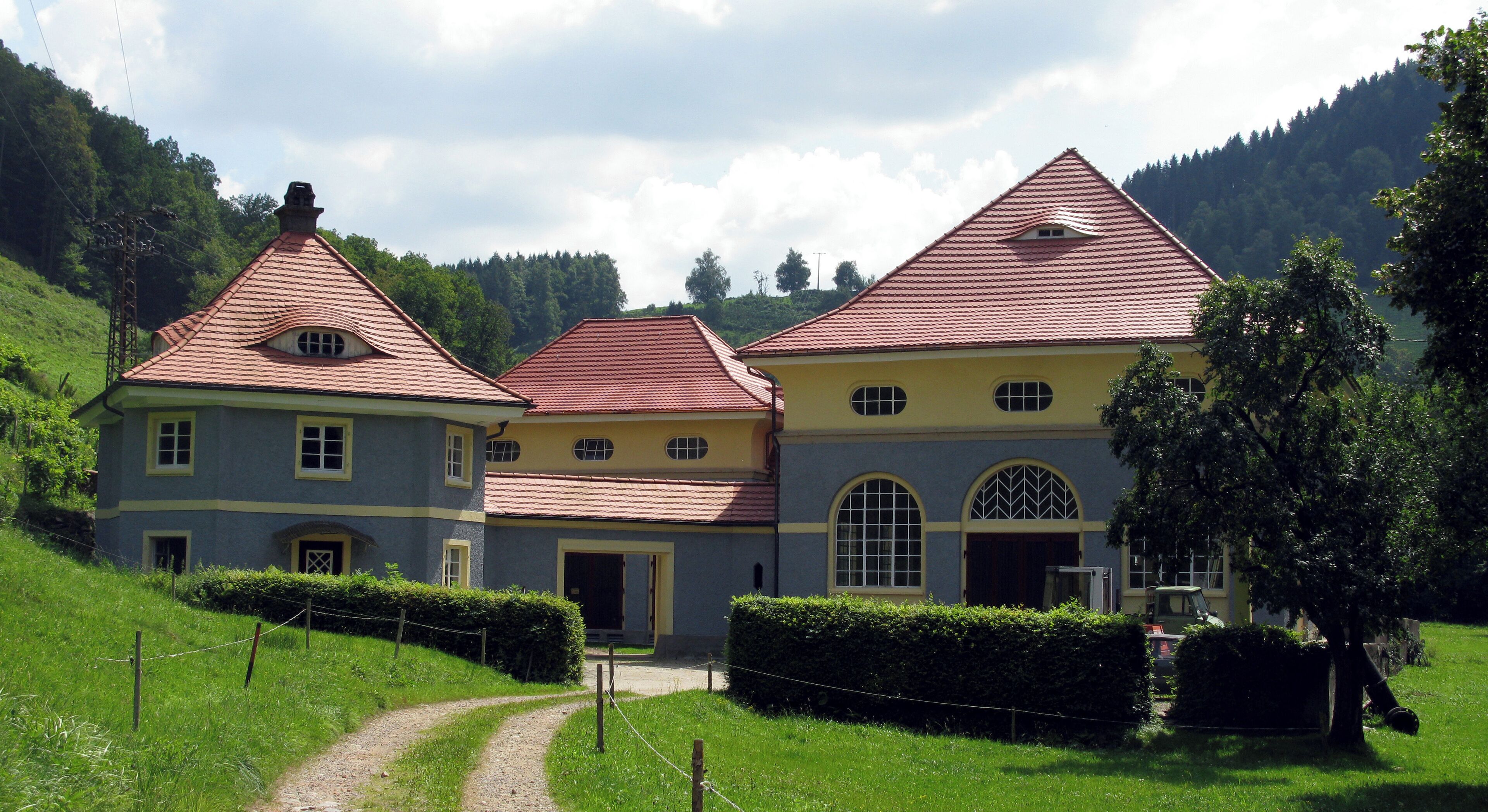 A large, multi-story building with a red-tiled roof, surrounded by lush greenery and hills.