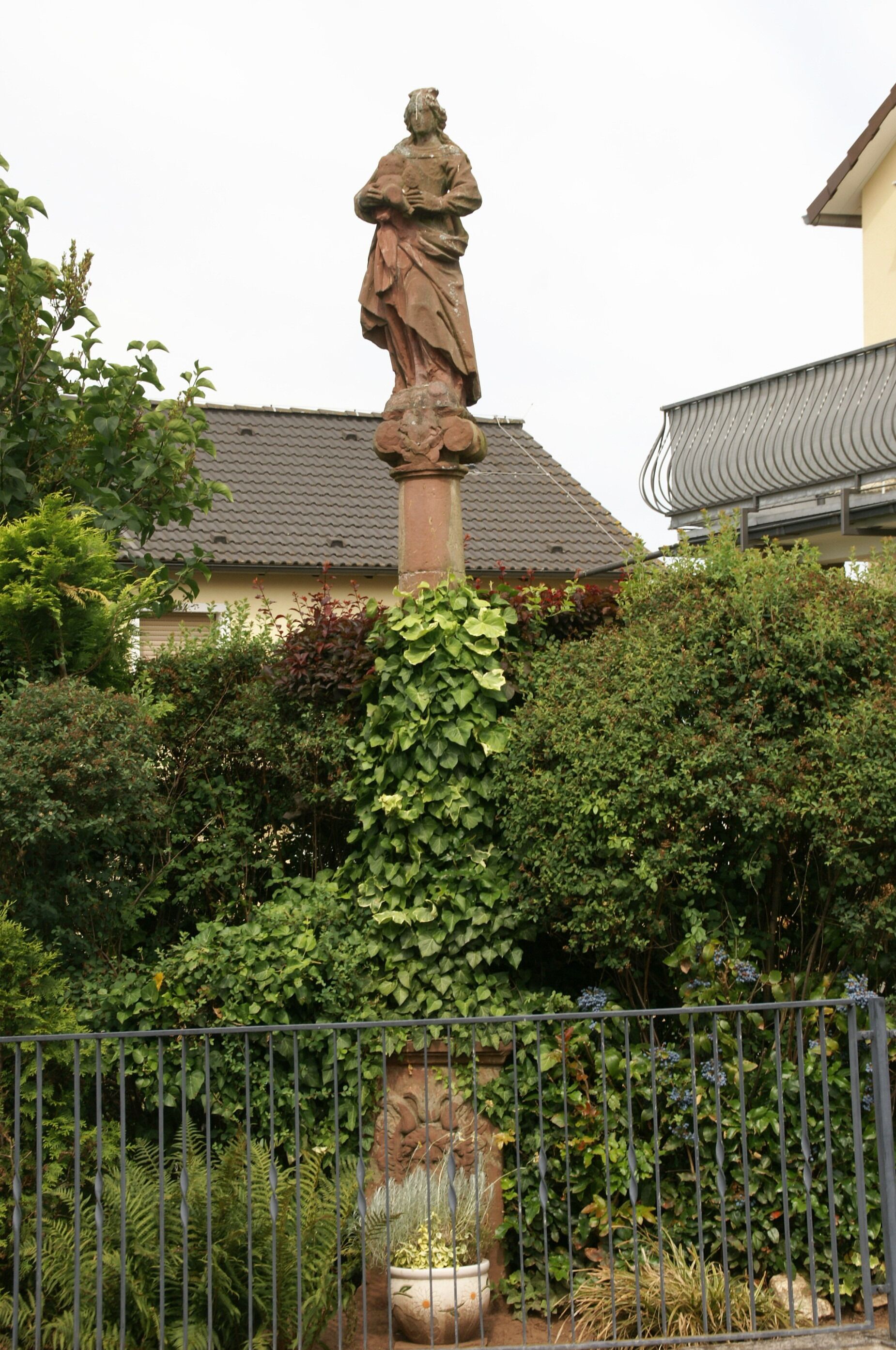 A stone statue in a garden with a fence, surrounded by lush greenery and a house in the background.