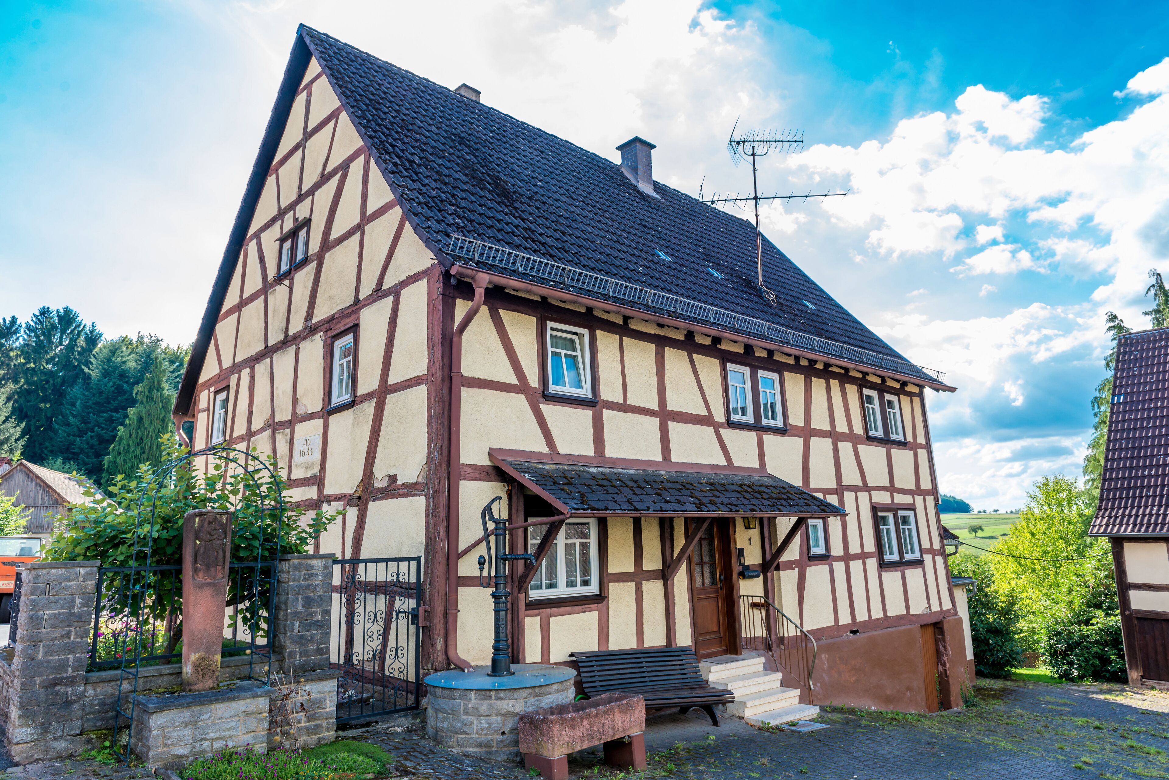 A traditional half-timbered house with a steeply pitched roof, a stone bench, and a wrought iron gate.