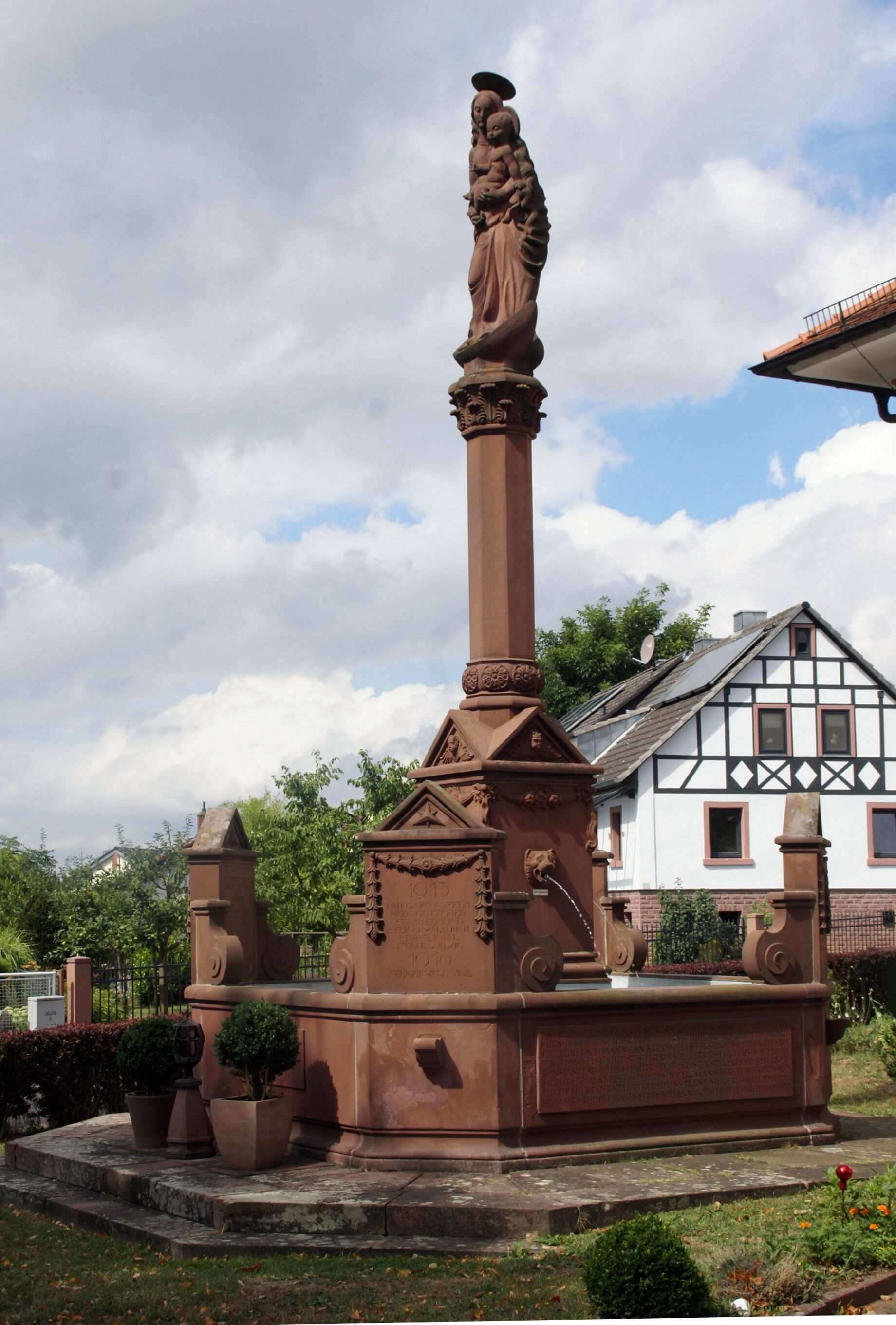 A tall, brown monument with a statue on top, surrounded by potted plants and a traditional house in the background.