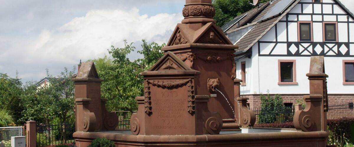A tall, brown monument with a statue on top, surrounded by potted plants and a traditional house in the background.