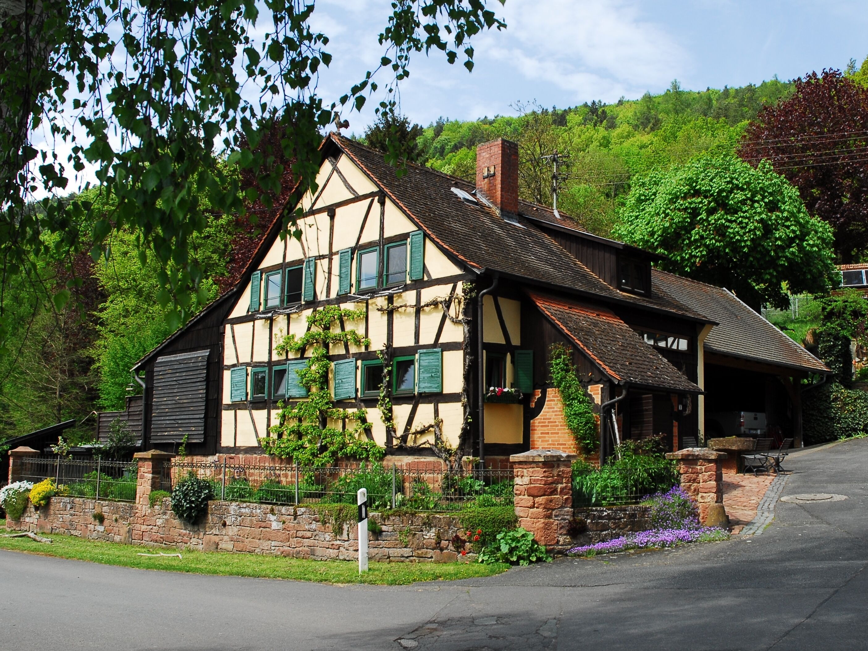A traditional half-timbered house with a sloped roof, surrounded by lush greenery and a stone wall.