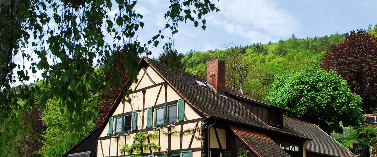 A traditional half-timbered house with a sloped roof, surrounded by lush greenery and a stone wall.