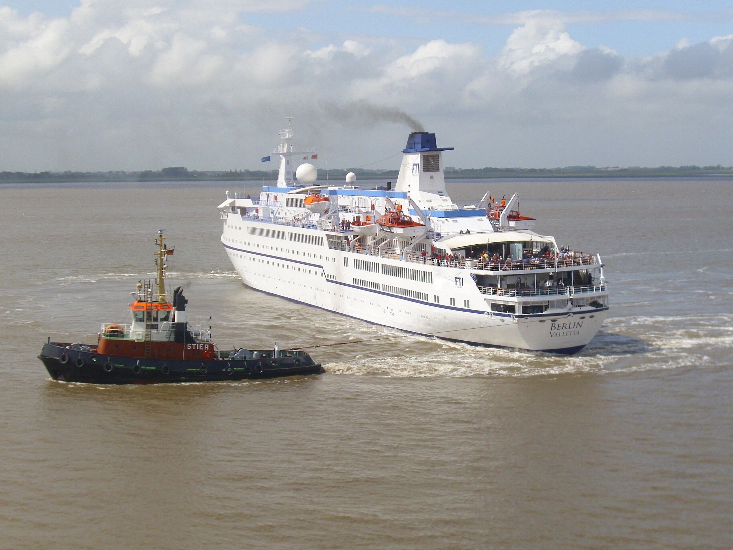 A large cruise ship named 'BERLIN VALLETTA' with a tugboat escorting it.