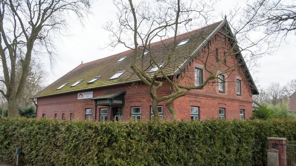 A red brick building with a steeply pitched roof, surrounded by a dense hedge.