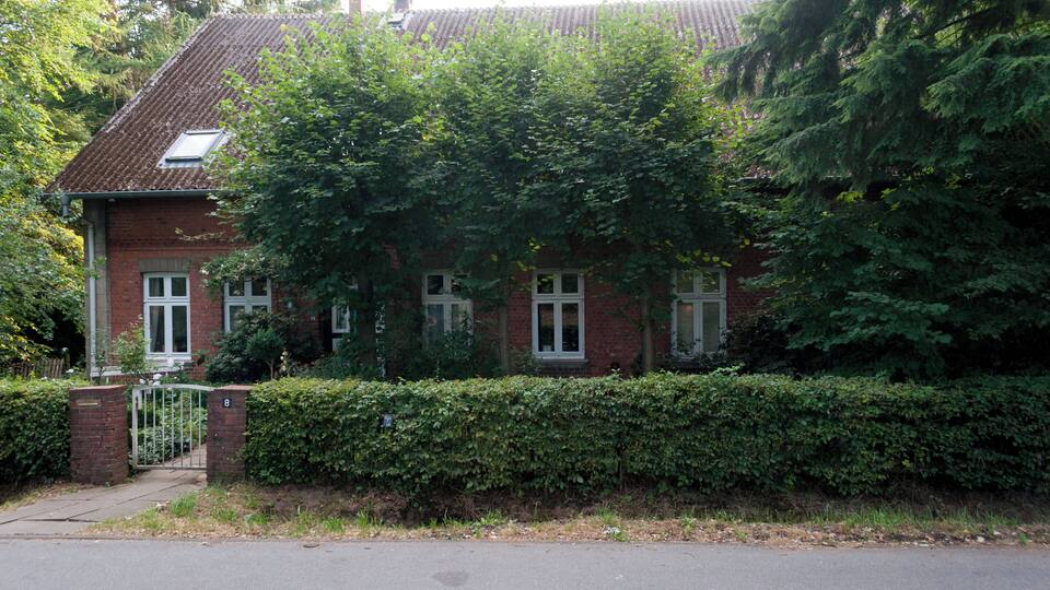 A red brick house with a tiled roof, surrounded by dense green foliage and a well-maintained hedge.