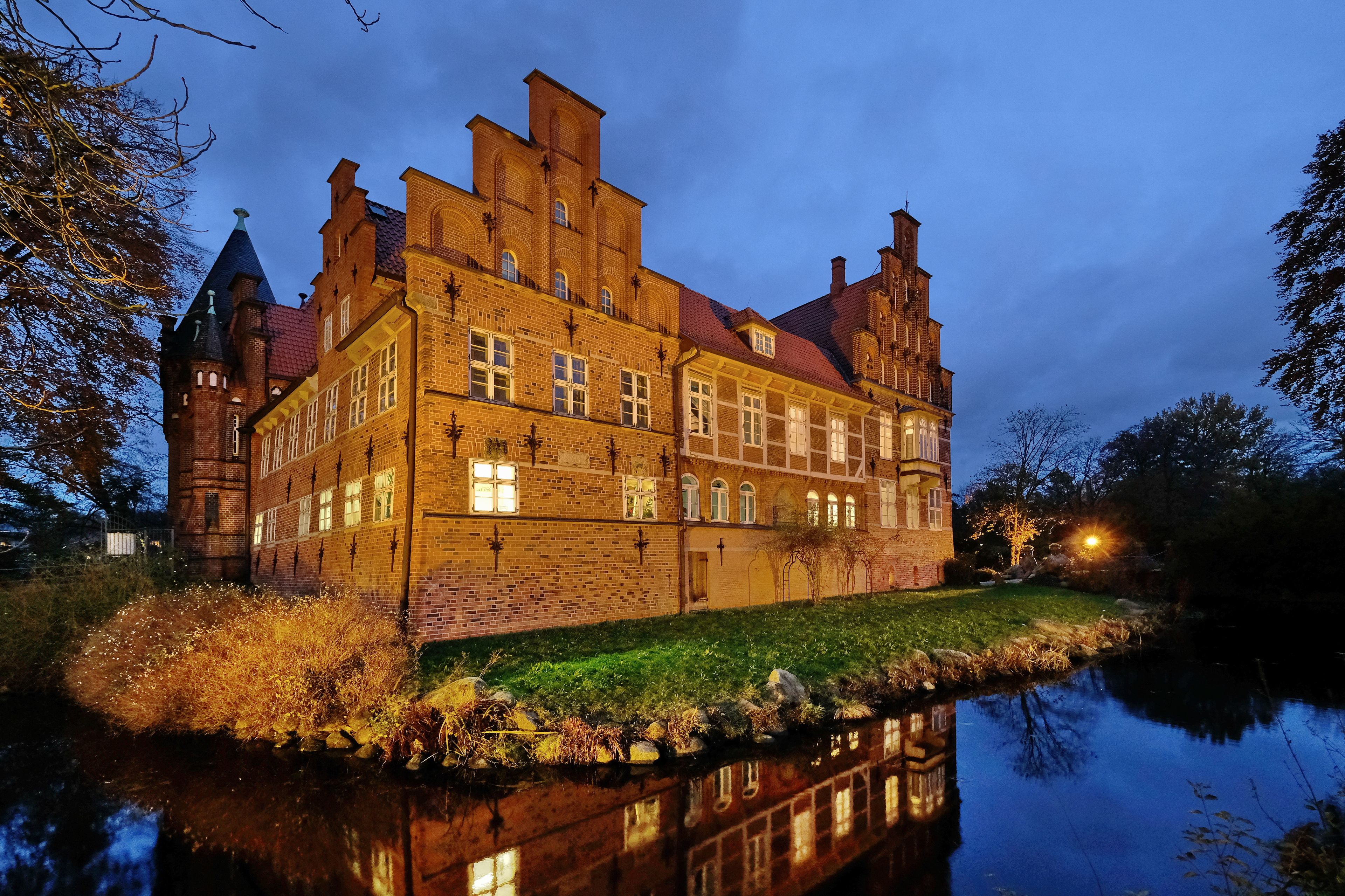 A large, historic brick building with multiple towers and a reflective water body in front.