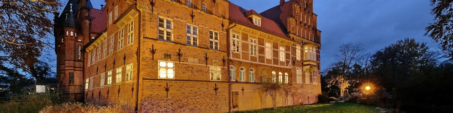 Um grande edifício histórico de tijolos, com várias torres e um corpo d'água refletiva à frente.