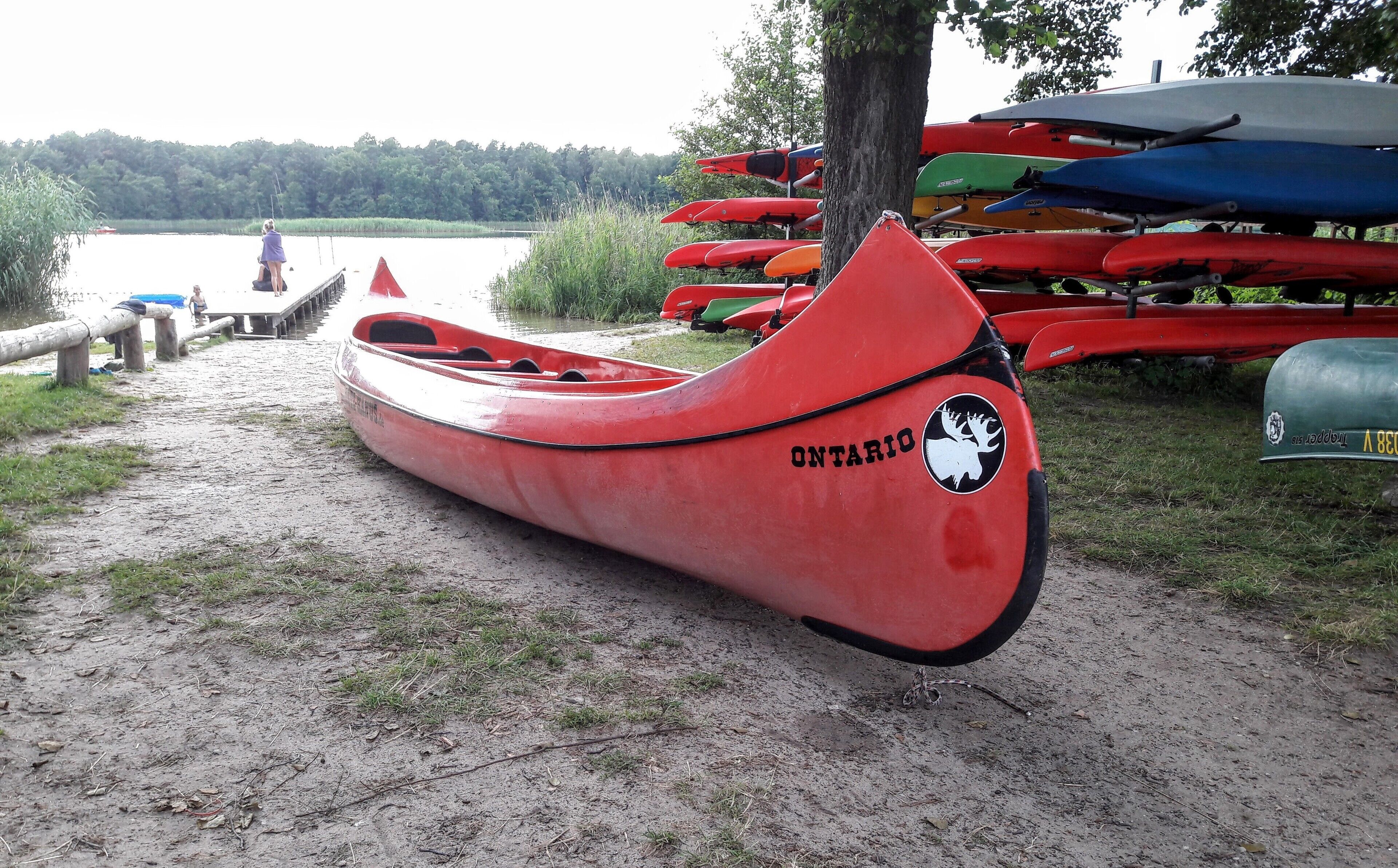 A red canoe with the word "ONTARIO" on it, parked on a dirt path near a lake with other canoes in the background.