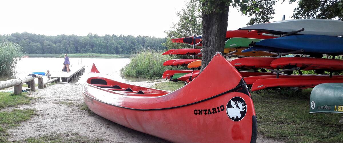 A red canoe with the word "ONTARIO" on it, parked on a dirt path near a lake with other canoes in the background.
