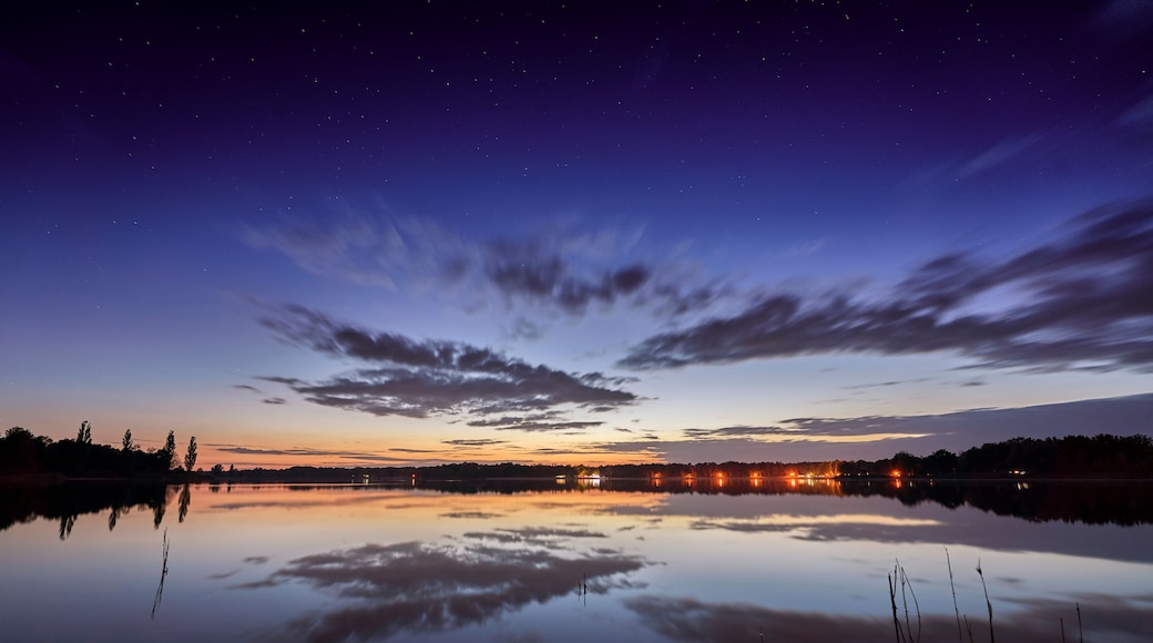 Ein Sternenhimmel, ein See, der den Himmel spiegelt, und in der Ferne Stadtlichter.