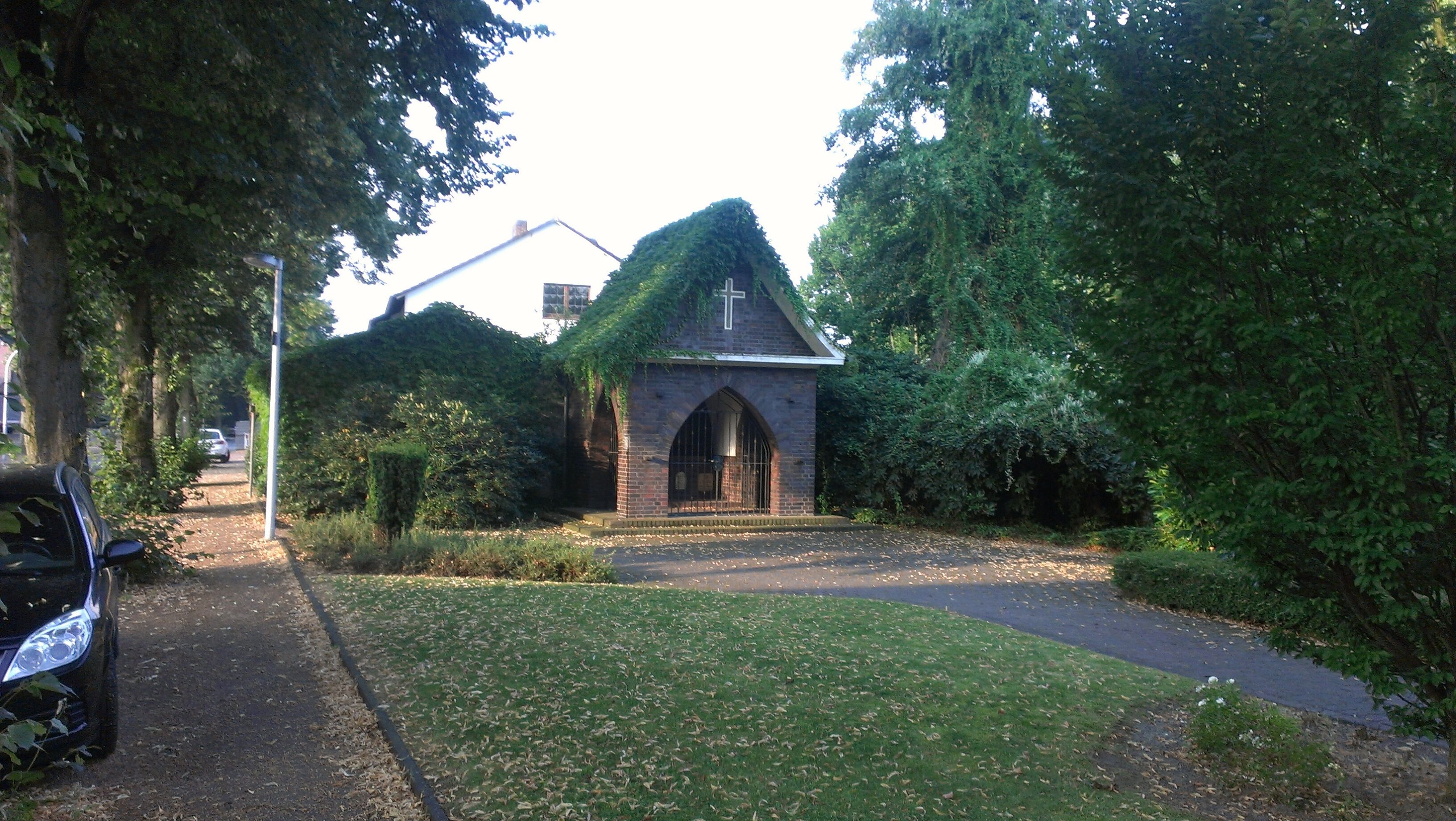 A small brick church with a cross on top, surrounded by trees and a paved path.