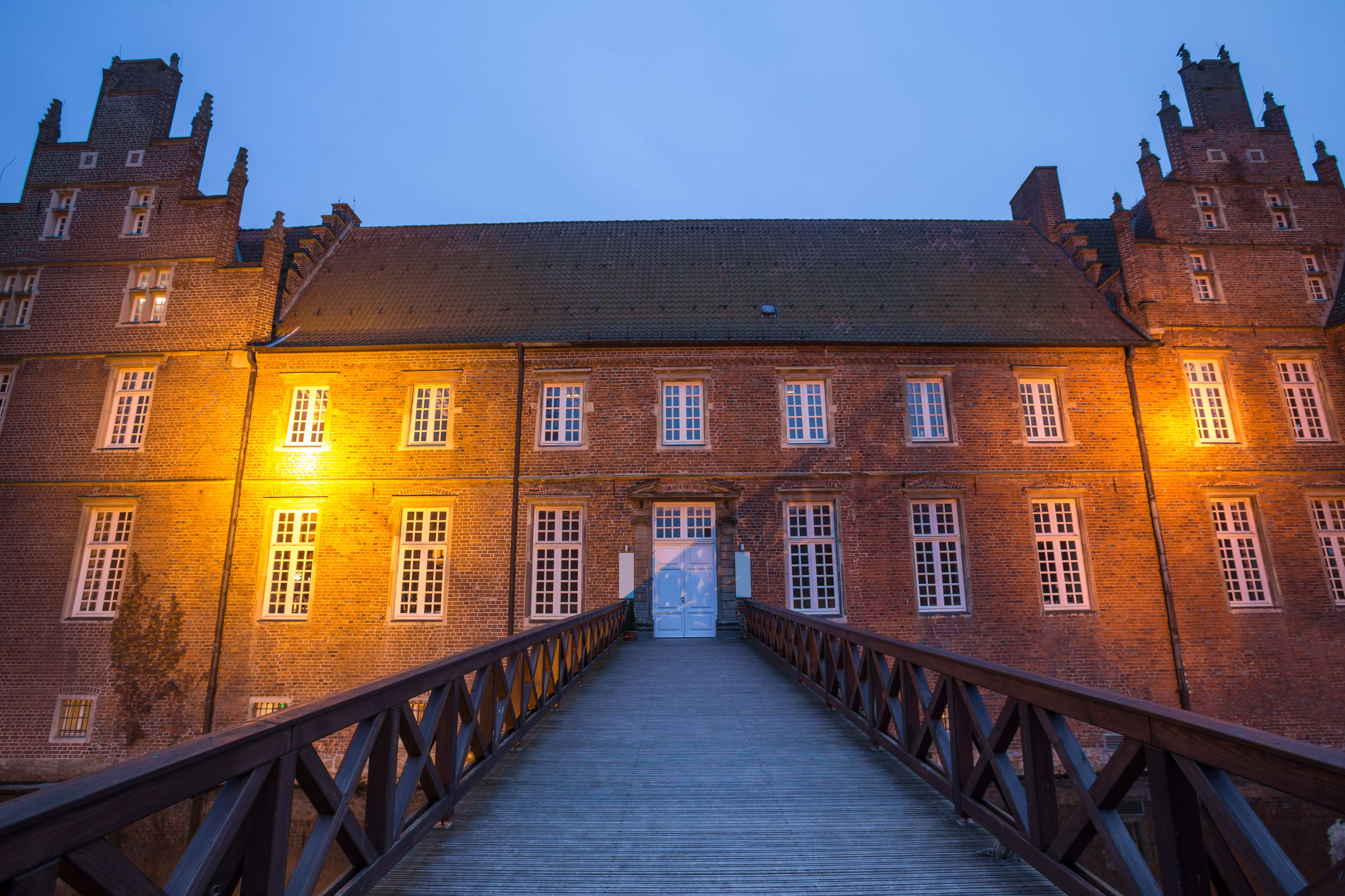 Eine Holzbrücke führt zu einem großen Backsteinhaus mit vielen Fenstern und einem zentralen Eingang.