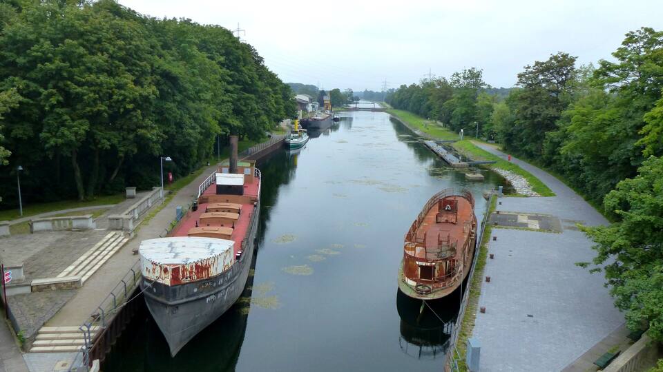A canal with two large cargo ships docked, surrounded by lush green trees and a clear sky.