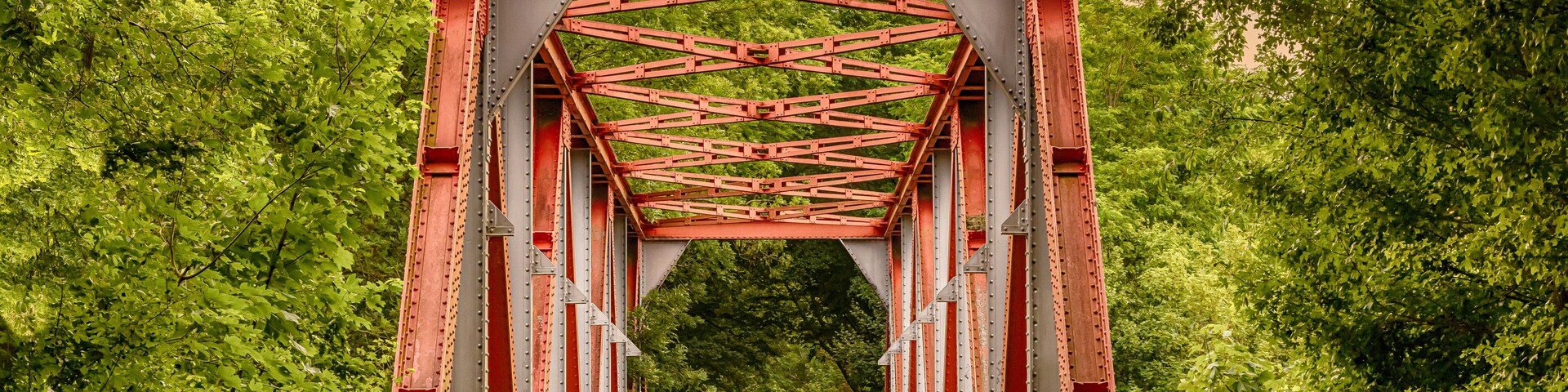Uma ponte de metal vermelha com design simétrico, rodeada por árvores verdes e exuberantes.